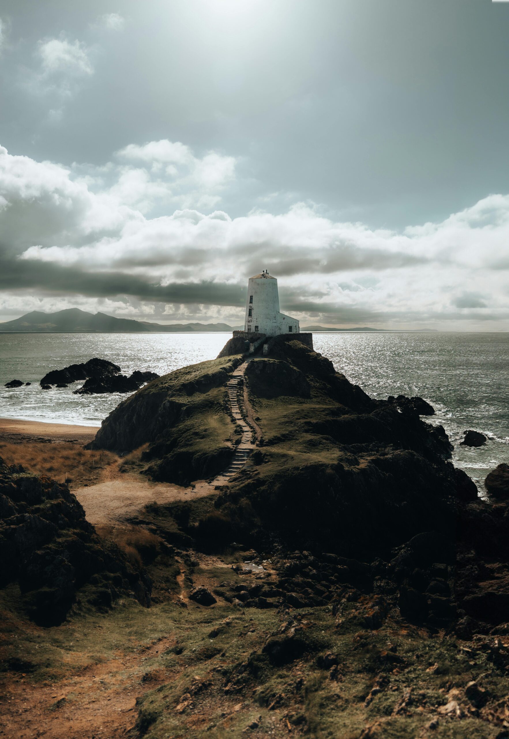 Scenic view of Twr Mawr Lighthouse atop a hill on Llanddwyn Island during sunset, surrounded by sea and coastline.